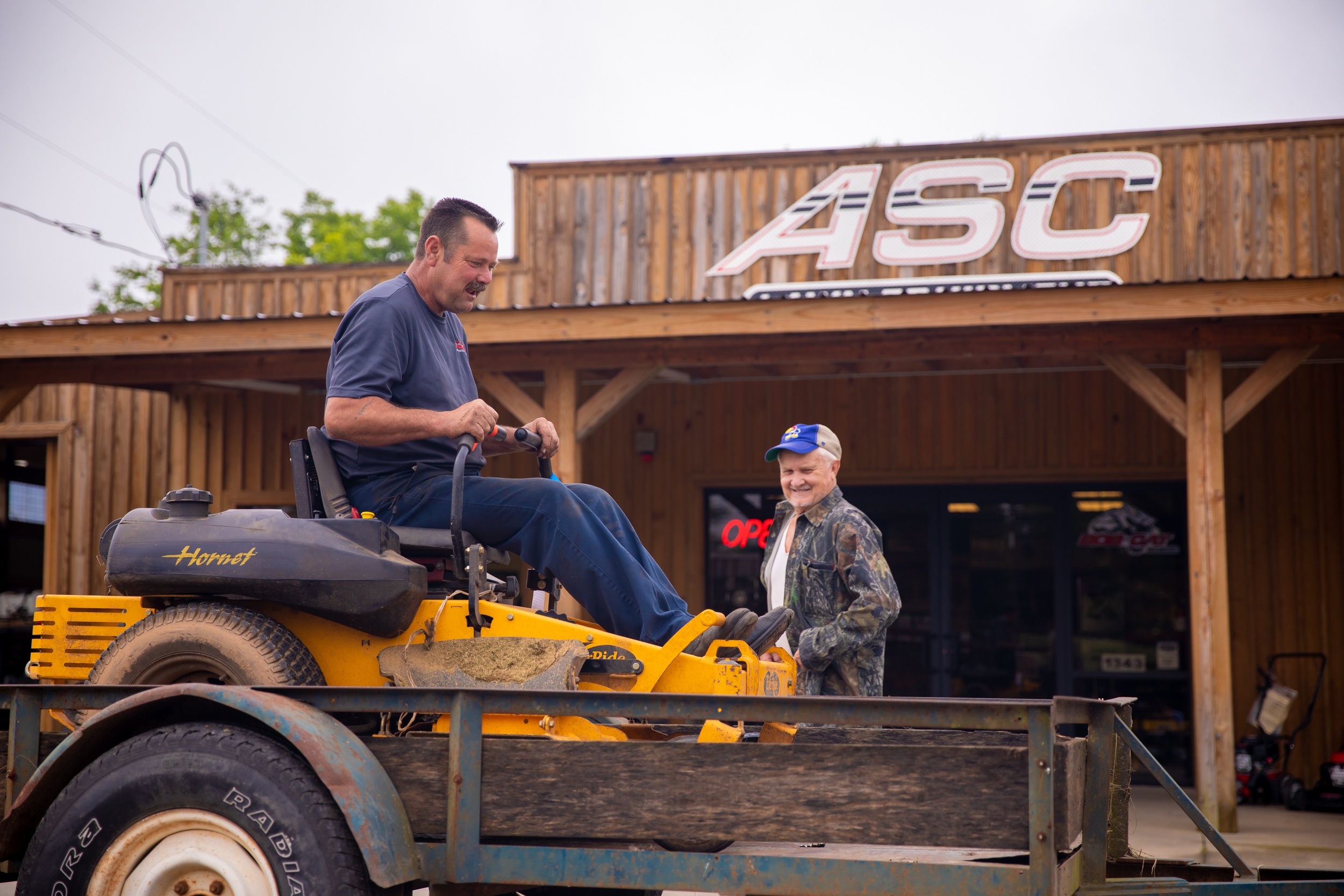 person driving a commercial lawn mower onto a trailer while another man looks on in front of the ASC Lawn Equipment building
