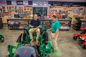 man sitting in a commercial lawn mower talking to a salesperson inside the store.