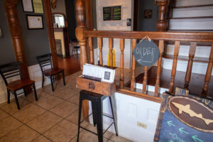 entrance of south of heaven bbq. hostess station with menus in a box on a stool. Chairs along the wall and a stair railing in the background.